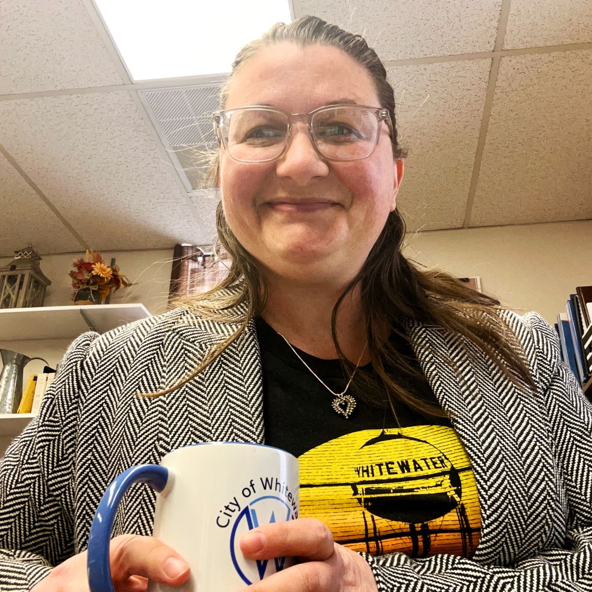 Brown haired woman with a jacket and glasses holding a coffee cup smiling at the camera